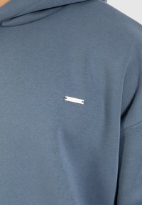 Felpa con cappuccio in morbido tessuto blu, dotata di un piccolo etichetta logo argento sul petto, con una finitura liscia e strutturata.