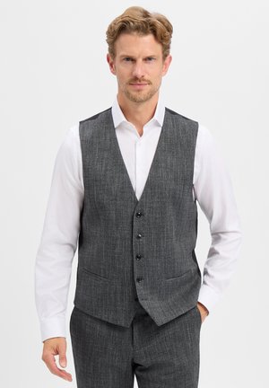 Man with short curly hair wearing a white shirt and gray buttoned vest and matching gray pants against a plain background.