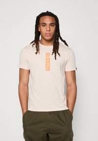 Lys beige t-shirt lavet af blødt stof med et orange lodret "ALPHA" logo i midten. Korte ærmer og rund halsudskæring.