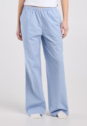 Weit geschnittene hellblaue Hosen mit Nadelstreifen, getragen von einer stehenden Person, kombiniert mit weißen Socken und einem kurzen weißen Oberteil.