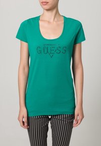 Camiseta de algodón verde con escote en forma de u, que presenta un gráfico de "GUESS" con pedrería en el centro. Mangas cortas y corte relajado.