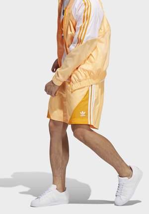 Chándal de satén naranja con acentos blancos, que presenta tres rayas blancas en las mangas y en los pantalones cortos. Zapatillas blancas completan el look.