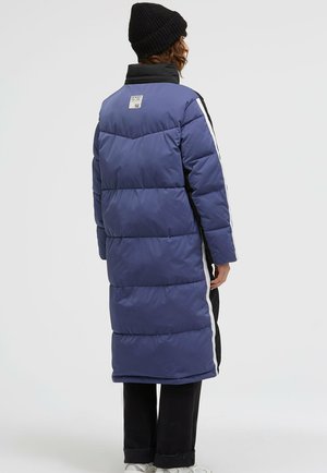 Long puffer coat in dark blue with horizontal stitching, black collar, and white side stripes. Worn over black pants and white sneakers.