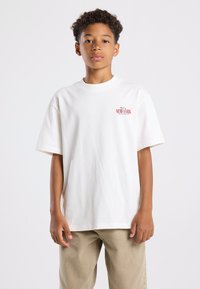 Valkoinen puuvilla t-paita lyhyillä hihoilla, pyöreällä kaula-aukoksi, jossa on pieni punainen "NEW YORK" -grafiikka vasemmassa rintakehässä.