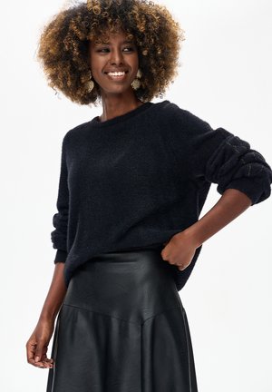 Uśmiechnięta kobieta z kręconymi włosami, nosząca duże kolczyki, czarny puszysty sweter i czarną skórzaną spódnicę na białym tle.
