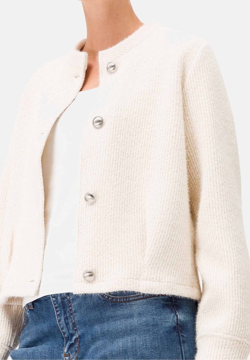 Cardigan en maille texturée couleur crème avec des boutons argentés, porté sur un haut blanc et associé à un jean en denim bleu.