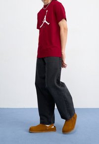 Czerwona bawełniana koszulka z białym logo, czarne szerokie jeansy oraz brązowe zamszowe chodaki z bocznym guzikiem, na tle jednolitej ściany.