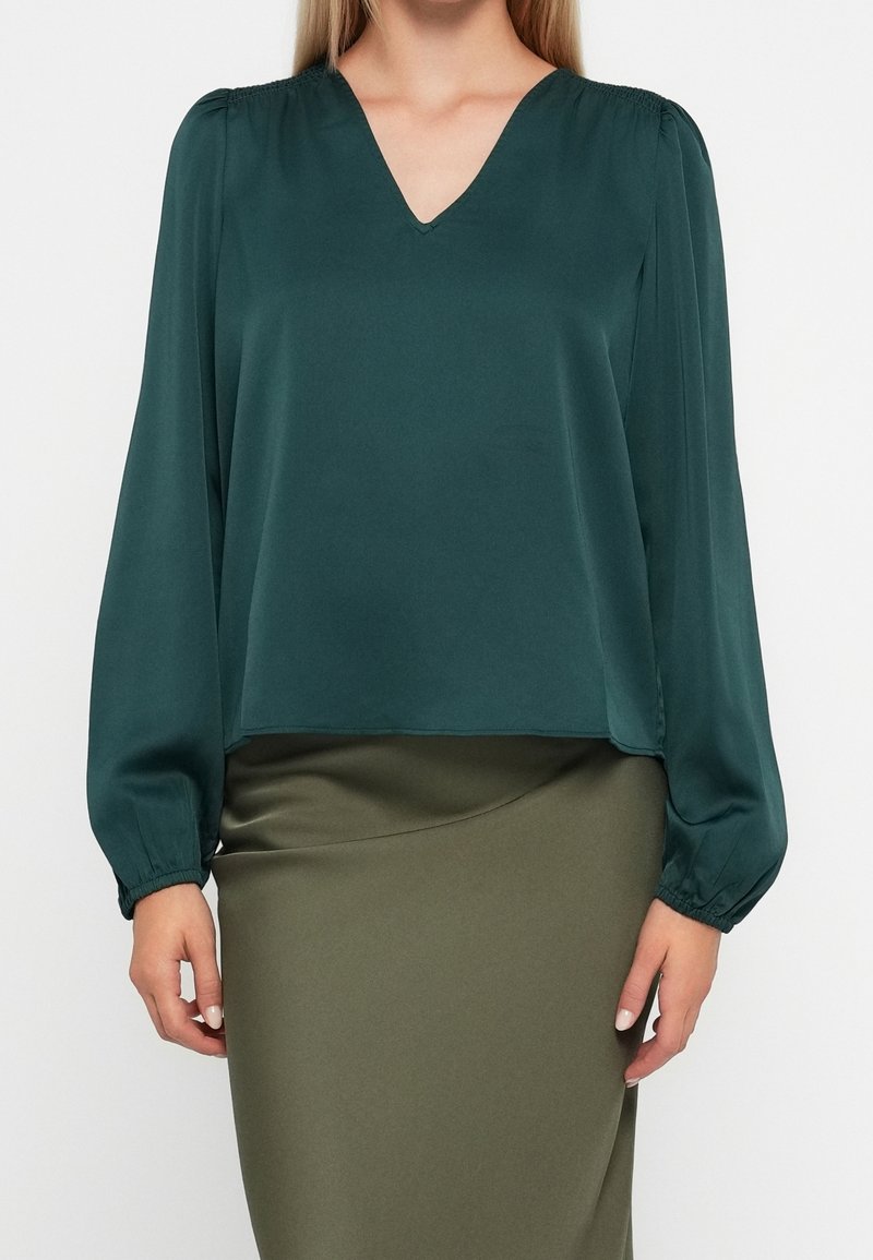 Femme portant un chemisier vert foncé à col en V et manches longues et une jupe vert olive, debout devant un fond uni.