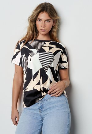 Frau mit langen hellbraunen Haaren, die ein schwarz-beiges T-Shirt mit abstraktem Muster und hellblaue Jeans trägt, posiert vor einer schlichten Wand.