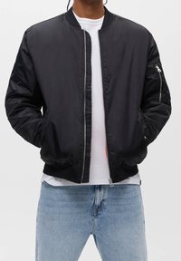 Veste bomber noire en tissu brillant, dotée d'une fermeture éclair, de poignets côtelés et d'une poche latérale avec fermeture éclair. Portée par-dessus une chemise blanche.