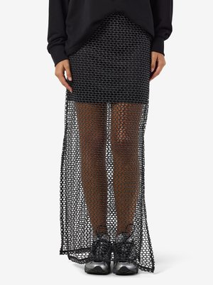 Žena v čiernej maxi sukni z priesvitnej sieťoviny cez minisukňu a čiernych teniskách, stojaca s uvoľnenými rukami pozdĺž tela na bielom pozadí.