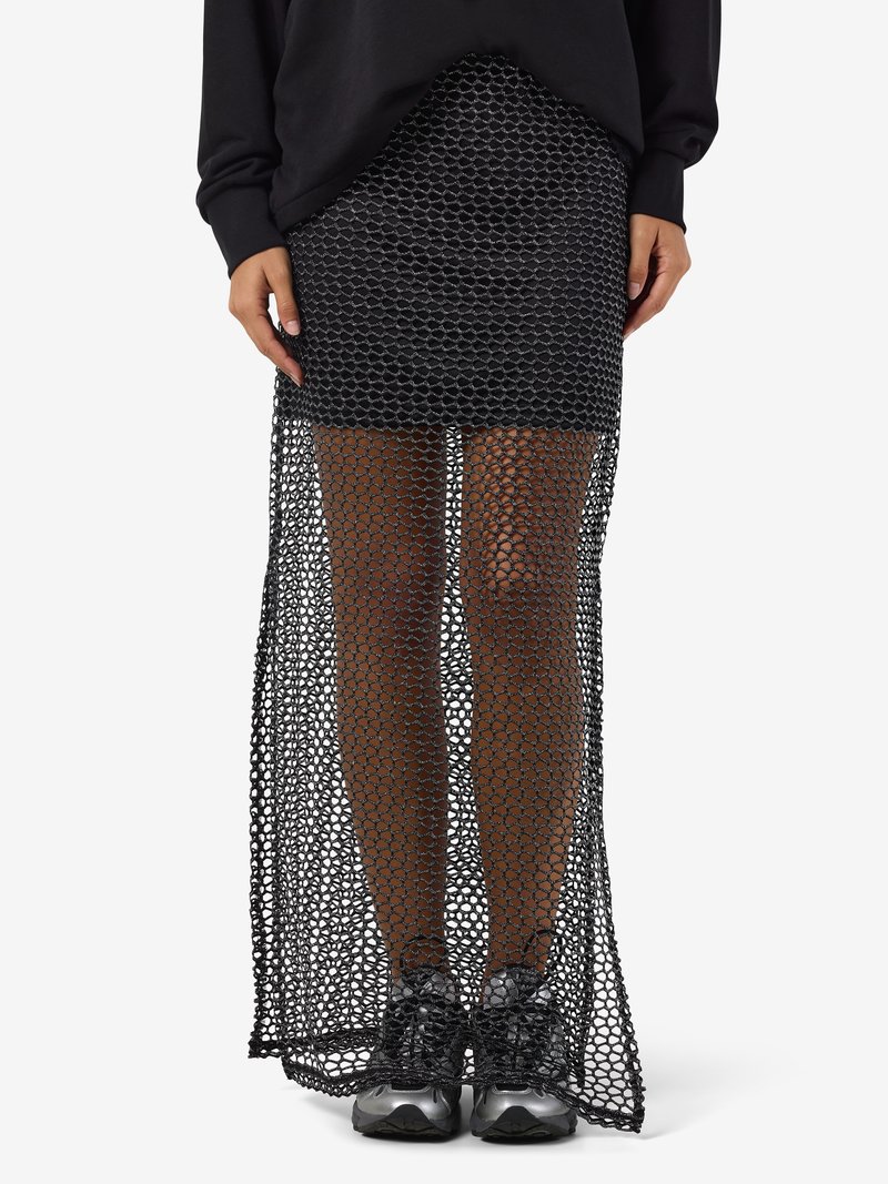 Γυναίκα που φοράει μαύρη μάλλινη μακριά φούστα πάνω από μια μίνι φούστα και μαύρα αθλητικά παπούτσια, στέκεται με τα χέρια χαλαρά στο πλάι σε λευκό φόντο.