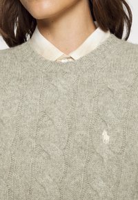 Gros plan sur une personne portant un pull gris clair en maille torsadée par-dessus une chemise crème boutonnée, avec un petit logo de cheval brodé sur le pull.