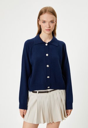 Tummansininen neulecardigan napilla edessä, jossa on ribbauskaulus ja hihansuut. Yhdistetty pleatattuun beiget hameeseen.