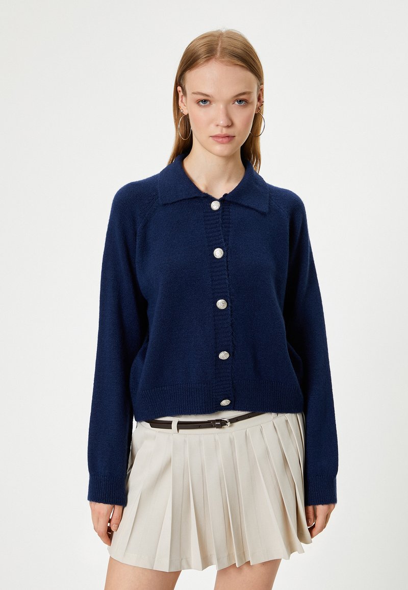 Marineblaues Strick-Cardigan mit Knopfleiste, ausgestattet mit einem gerippten Kragen und Bündchen. Kombination mit einem plissierten beigen Rock.