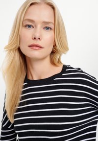 Czarny i biały sweter z prążkowanym wzorem, o okrągłym dekolcie, z gładką fakturą. Modelka ma proste, długie blond włosy.