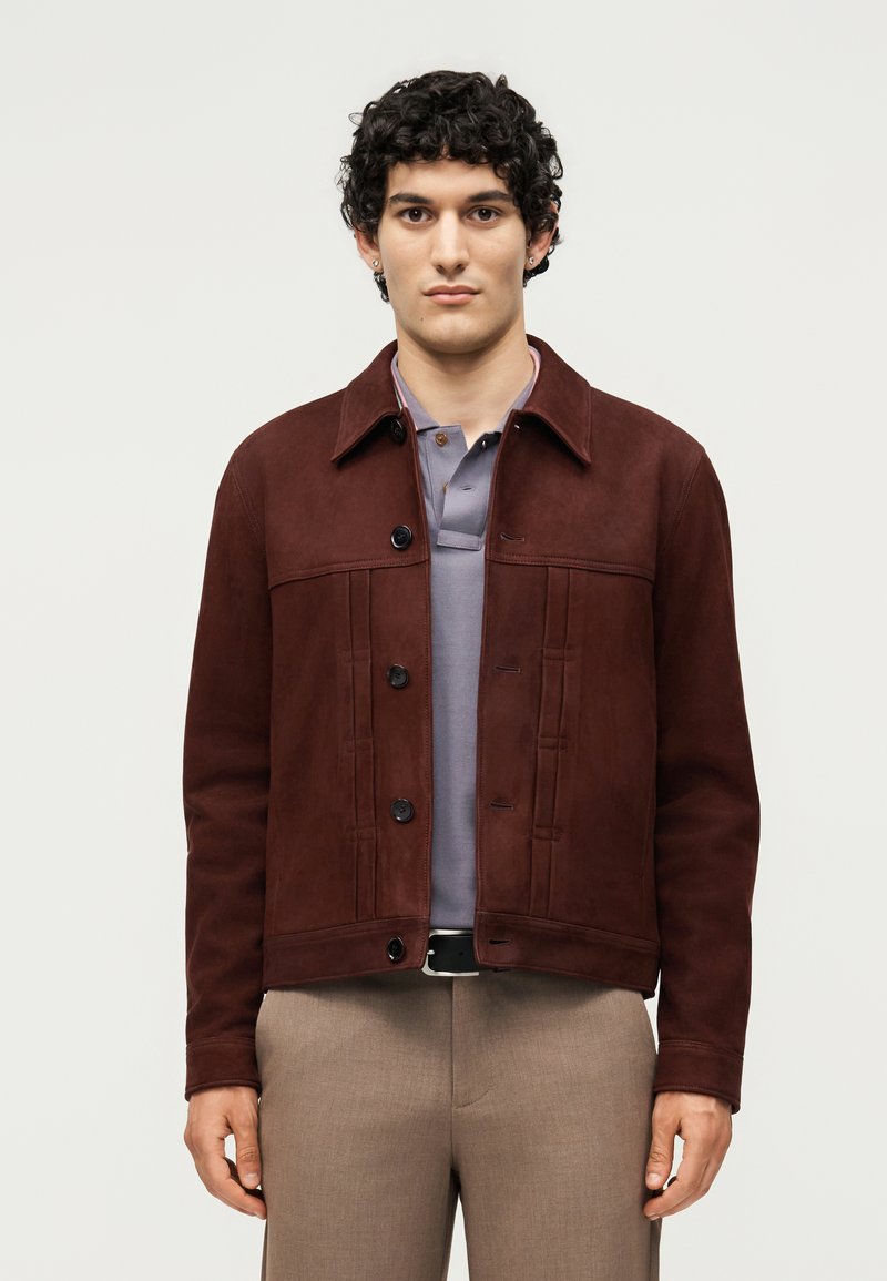 Άνδρας με σγουρά μαλλιά φοράει σκούρο καφέ κουμπωτό σακάκι πάνω από γκρι πόλο πουκάμισο και ανοιχτό καφέ παντελόνι σε απλό φόντο.