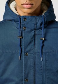 Chaqueta azul marino con un forro suave y de color claro. Presenta botones metálicos, capucha con cordón y detalles de costura.