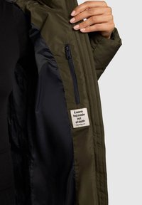 Olivengrøn isoleret jakke med sort foer. Har en lynlåslomme og et stofmærke med tekst. Glat yderside med quiltet design.