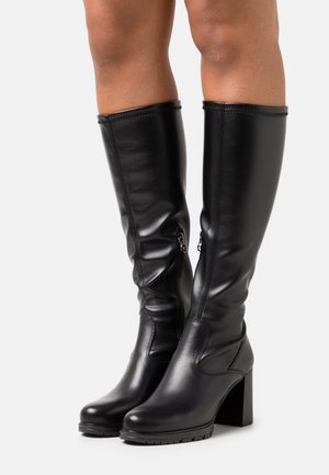 Czarne skórzane oficerki do kolan na obcasach typu słupek, z bocznymi zamkami i zaokrąglonymi czubkami, noszone na gołych nogach na jasnym tle.