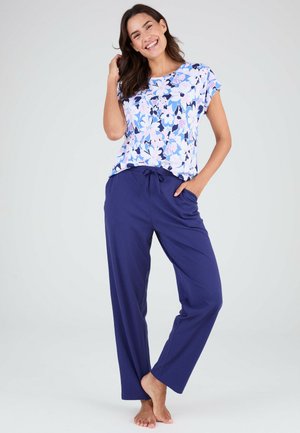 Lachende vrouw staat blootsvoets met hand in zak, gekleed in een blauw bloesje met korte mouwen en effen marineblauwe losse broek.