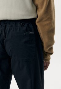 Person iført sorte Columbia-bukser med baglomme og beige og brunt langærmet top, vist fra taljen og ned mod hvid baggrund.