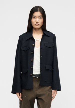 Azjatka ubrana w czarną kurtkę zapinaną na guziki, na białą bluzkę, w połączeniu z brązowymi spodniami i czarnym paskiem z srebrnymi ćwiekami.