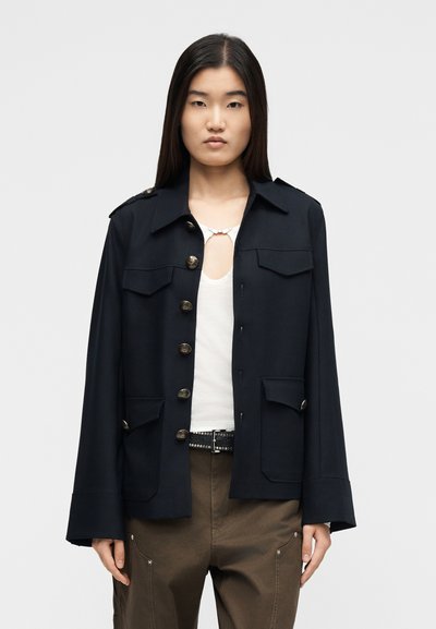 Femme asiatique portant une veste noire boutonnée sur un haut blanc, associée à un pantalon marron et une ceinture noire à clous argentés.
