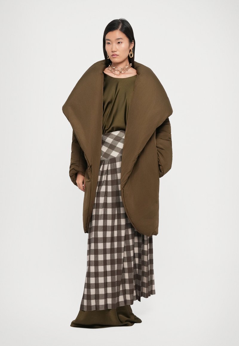 Abrigo de plumas oversized en verde oliva, falda larga a cuadros con cuadrados grises y blancos, superpuesta a una blusa de satén. Decorada con joyería de metal.
