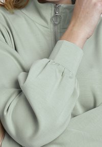 Parte superior de color verde claro con cremallera, que presenta un cuello alto, mangas largas fruncidas y tela lisa y texturizada. La cremallera incluye un tirador circular.