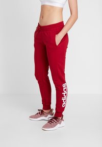 Röda joggerbyxor med vitt "adidas"-logotyp längs vänster ben, tillverkade av mjukt material, med elastisk midja och justerbar dragsko.