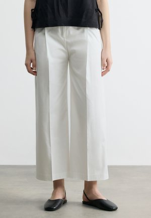 Osoba oblečená v bielych širokých 3/4 nohaviciach s prednými záhybmi, čiernom tielku bez rukávov a čiernych balerínach so zapínaním vzadu, stojaca na sivej podlahe.