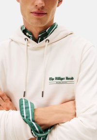 Joven con una sudadera blanca sobre una camisa de botones a rayas verdes y blancas, con los brazos cruzados. El texto en la sudadera dice "The Hilfiger Herald MCMLXXXV."