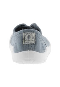 Zapato slip-on de lona gris con suela de goma blanca y una etiqueta en el talón. El diseño presenta un perfil bajo en la parte posterior y una textura suave.