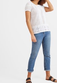 Haut blanc en œillet avec manches courtes, motifs floraux texturés, associé à un jean bleu clair retroussé et des sandales marron. Tenue simple et décontractée.