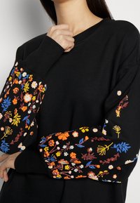 Sudadera negra con mangas largas, con intrincados bordados florales multicolores en naranja, azul y blanco. Textura suave, diseño informal.