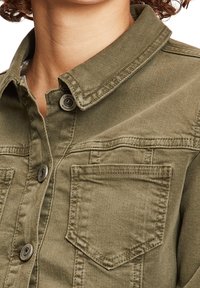 Camisa vaquera de color verde oliva con cuello, botones frontales y dos bolsillos en el pecho. Presenta costuras resistentes y tela texturizada.