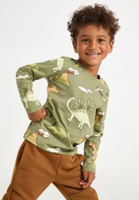 Camicia verde a maniche lunghe con stampe di dinosauro e vulcano, abbinata a pantaloni marroni. Tessuto di cotone con texture liscia.