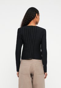 Kvinde med langt krøllet hår iført en tætsiddende sort tekstureret langærmet top og beige højtaljede bukser, vist fra baghovedet.