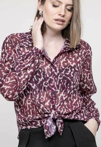 Blouse transparente bordeaux avec un motif léopard ; possède des manches longues, une fermeture à boutons et un détail de ourlet noué.