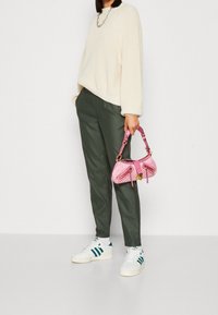 Pull en maille crème, pantalon vert olive, baskets blanches avec des rayures bleues, tenant un petit sac à main rose avec des détails en zip et des anses contrastantes.
