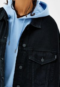 Svart denimjacka med knäppning och fickor, lagrad över en ljusblå hoodie med dragsnören. Mjuk tygstruktur synlig.