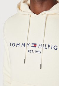 Kermainen huppari, joka on valmistettu pehmeästä kangasta ja jossa on brodeerattu logo tummansinisellä ja punaisella. Sisältää narut ja metalliset kärjet.