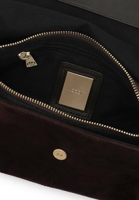 Sötétbarna suede táska belső része arany cipzárral, fekete szövet béléssel és arany logolemezzel rendelkezik. A mágneses patent zár is látható.