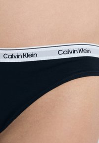 Čierne bavlnené bikiny s bielym elastickým pásom, na ktorom je čiernymi písmenami napísané "Calvin Klein". Mäkkučká textúra a hladký povrch.