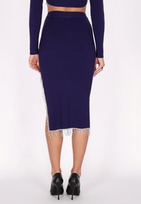 Jupe midi en tricot violet avec un ourlet à franges orné de perles en argent. Design ajusté, texture côtelée et détail fendu sur le côté. Associée à des talons noirs.