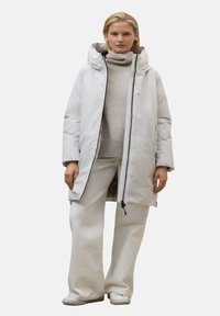 Abrigo acolchado blanco con capucha, con cierre de cremallera y bolsillos laterales. Se lleva sobre un suéter gris claro y pantalones blancos de pierna ancha.