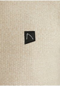Tissu beige texturé avec une petite étiquette noire en forme de trapèze comportant un design minimaliste à lignes blanches au centre.