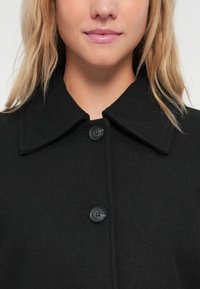Svartullskappa med en stor krage, två svarta knappar och en slät yta, visad på en person från bröstet och upp.