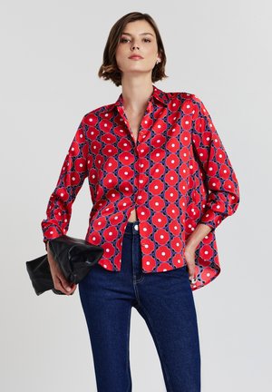 Camicia a maniche lunghe a motivi rossi con grandi disegni floreali circolari, abbinata a jeans blu scuro e una borsa a portafoglio nera. Tessuto liscio, vestibilità comoda.